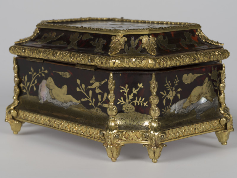 zbliżenie na dekorację prawego tylnego narożnika, widok z boku, motyw roślinny po środku, po prawej stronie śpiący amorek, motywy roślinne, po lewej leżący amorek i lecący ptak,  krawędzie szkatułki pokryte dekoracjami z brązu w postaci palmet i ornamentu kwiatowego, nóżki szkatułki w postaci rozety liści.