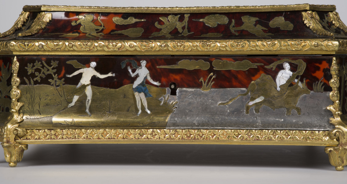 zbliżenie na inkrustowaną dekorację przedniej ścianki przedstawiającą Porwanie Europy, widok z boku na ściankę boczną szkatułki. Po prawej kobieta jadąca na byku po lewej kobiety i mężczyzny. Krawędzie szkatułki pokryte dekoracjami z brązu w postaci palmet i ornamentu kwiatowego, nóżki szkatułki w postaci rozety liści.