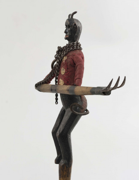 Figurka z szopki, diabeł, rzeźbiona w drewnie. Pomalowana na czarno. Ubrana w czerwony kaftan. Na szyi łańcuch. Do łańcucha doczepione widły, które diabeł trzyma obydwoma rękami. Twarz mocno modelowana, głębokie oczodoły, duże uszy, mała broda wysunięta mocno do przodu. Na głowie małe różki. Z tyłu skórzany ogon.