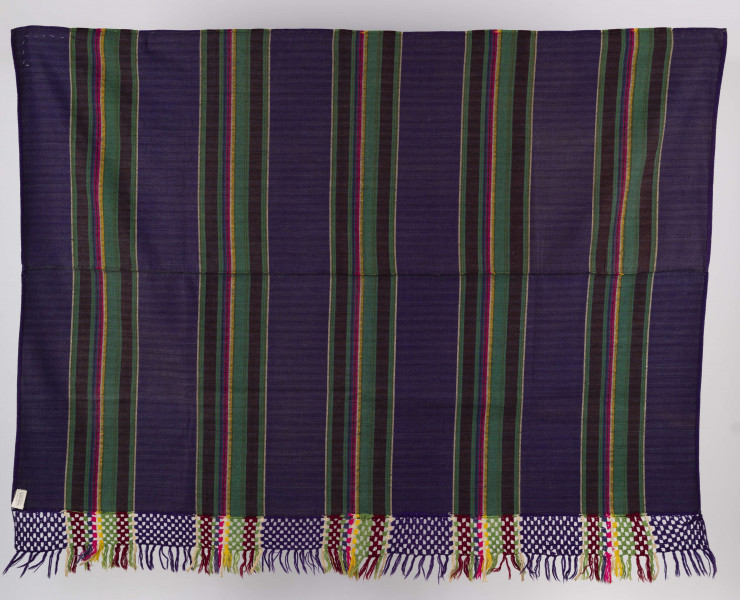 E/6045/ML - Tkanina pasiak tkana, zszyta z 2 części pośrodku. Splot rządkowym. Osnowa lniana czarna, wątek wełniany w kolorach: fioletowy, zielony, brązowy, żółty, amarantowy, biały, ciemnoniebieski. Raport tworzą poprzeczne wiązki pasów oddzielone szerokimi pasami w kolorze fioletowym. Jedna z krawędzi wzdłużnych wykończona szydełkową koronką z wełny w kolorach występujących w tkaninie. Zakończona wiązanymi frędzlami. 