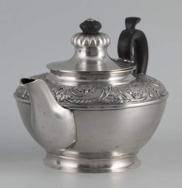 S/Mt/542/ML - Korpus w kształcie spłaszczonej kuli, na niskiej stopie, wylew w formie dzióbka, ucho drewniane malowane na czarno, osadzone w dwóch tulejkach, pokrywka na zawiasie, płaska, w części środkowej wypiętrzona i ozdobiona puklowaną rozetą.Górna część brzuśca ozdobiona sztancowaną taśmą z wzorem spiralnie zwinietej wici roślinnej z kwiatami, uchwyt przykrywki zakończony drewnianą rozetką.  
