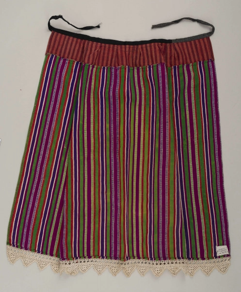 E/8132/ML - Fartuch wełniany szyty maszynowo. Wykonany z materiału samodziałowego tkanego w pionowe paski, w kolorach: czerwonym, białym, szafirowym, bordowym, zielonym. Uszyta z prostokątnego kawałka materiału, do którego doszyto z obu stron kliny. U góry fartuszek przedłużony przez wszycie kawałka materiału szer. 7 cm, w pasy czerwono-granatowe. W talii wszyty w pasek z czarnego materiału, szer. 2 cm, zakończony troczkami. Dół fartuszka zdobi biała koronka szer. 5 cm.