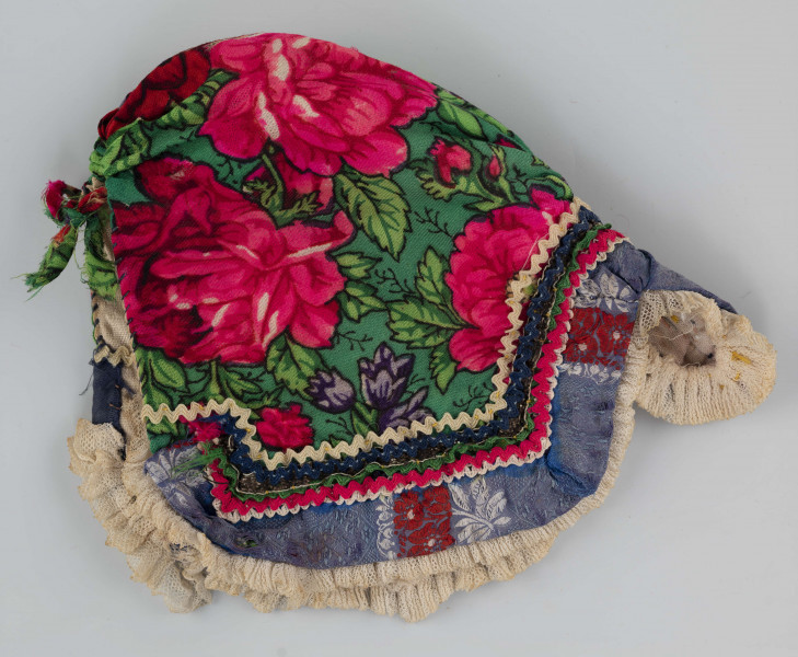 E/8141/ML - Czepek mężatki na płóciennej podszewce z zielonej cienkiej wełenki w amarantowe róże, fioletowe sasanki i jasnozielone listki. Marszczony na środku przodu i tyłu. Przód ozdobionymi kremowymi i granatowymi ząbkami, srebrną metaliczną wstążką oraz szeroką atłasową wstążką w kolorze granatowym w białe i czerwone kwiaty. Brzeg wykończony białą tiulową, drobno plisowaną kryzą. Przód wycięty w duży ząb w kształt płatka kwiatu.