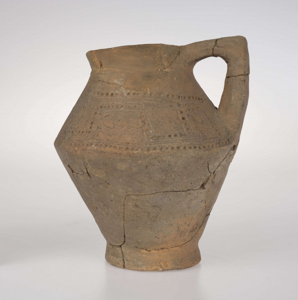 Dzban ostro profilowany, z prostym, odgiętym na zewnatrz brzegiem podkreślonym rowkiem, ostro zaznaczonym załomem brzuśca i wyodrębioną stopką. Ucho dzbana jest taśmowate, kolankowato zgięte, z przewierconym kanalikiem. Ponad załomem brzuśca naczynie zdobione jest ornamentem blankowym, który tworzy para równolegle rytych linii wypełnionych dołkami. Całość ujęta górą i dołem w wątek linii rytej i rzędu dołków. 
