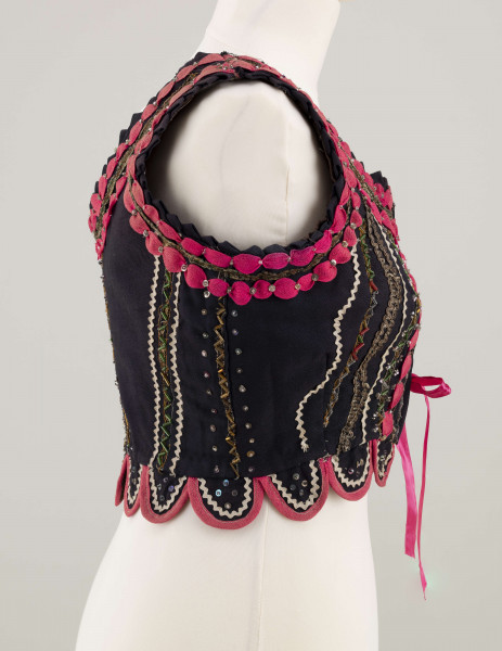 E/7358/ML - Gorset zszyty z 3 kawałków granatowej wełenki, podszyty brązową bawełnianą podszewką. Z przodu rozcięcie usztywnione fiszbinami, zaopatrzone w 10 haftek drucianych do przewlekania wstążeczki. Od pasa gorsetu dookoła opadają klapki. Cały gorset bogato zdobiony amarantowymi tasiemkami, żółtym i srebrnym szychem, białą krepiną, barwnymi koralikami i cekinami. Przód gorsetu, brzegi wykroju i pachy ozdobione dodatkowo granatowymi ząbkami. 