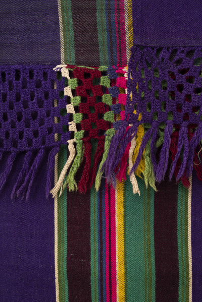 E/6045/ML - Tkanina pasiak tkana, zszyta z 2 części pośrodku. Splot rządkowym. Osnowa lniana czarna, wątek wełniany w kolorach: fioletowy, zielony, brązowy, żółty, amarantowy, biały, ciemnoniebieski. Raport tworzą poprzeczne wiązki pasów oddzielone szerokimi pasami w kolorze fioletowym. Jedna z krawędzi wzdłużnych wykończona szydełkową koronką z wełny w kolorach występujących w tkaninie. Zakończona wiązanymi frędzlami. 