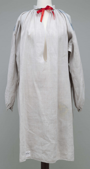 E/843/ML - Koszula kobieca o kroju przyramkowym, wykonana z szarego lnianego płótna. Długa, z niewielkim wykładanym kołnierzem z rozcięciem z przodu. Na przyramkach ozdobiona niebieskim haftem łańcuszkowym we wzory wolutowe, a na mankietach kolorową stebnówką. Noszona do stroju świątecznego. 