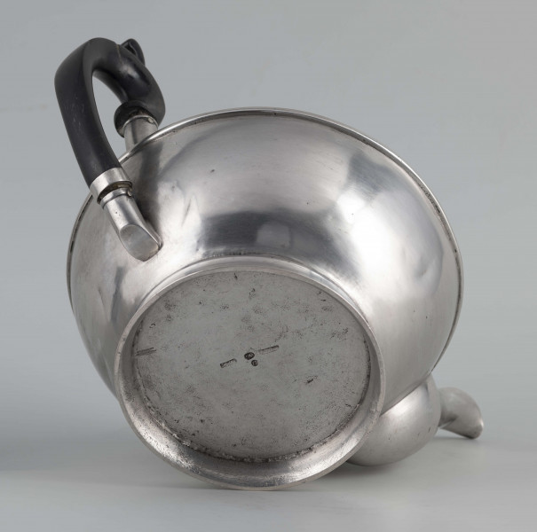 S/Mt/542/ML - Korpus w kształcie spłaszczonej kuli, na niskiej stopie, wylew w formie dzióbka, ucho drewniane malowane na czarno, osadzone w dwóch tulejkach, pokrywka na zawiasie, płaska, w części środkowej wypiętrzona i ozdobiona puklowaną rozetą.Górna część brzuśca ozdobiona sztancowaną taśmą z wzorem spiralnie zwinietej wici roślinnej z kwiatami, uchwyt przykrywki zakończony drewnianą rozetką.  
