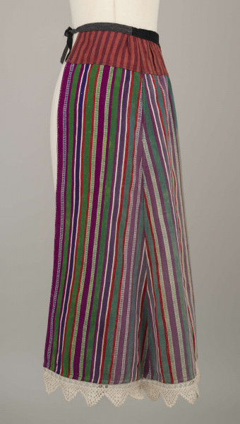 E/8132/ML - Fartuch wełniany szyty maszynowo. Wykonany z materiału samodziałowego tkanego w pionowe paski, w kolorach: czerwonym, białym, szafirowym, bordowym, zielonym. Uszyta z prostokątnego kawałka materiału, do którego doszyto z obu stron kliny. U góry fartuszek przedłużony przez wszycie kawałka materiału szer. 7 cm, w pasy czerwono-granatowe. W talii wszyty w pasek z czarnego materiału, szer. 2 cm, zakończony troczkami. Dół fartuszka zdobi biała koronka szer. 5 cm.