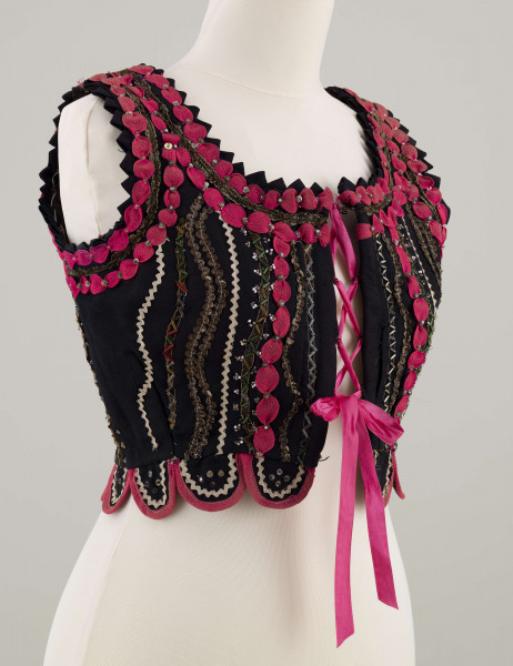 E/7358/ML - Gorset zszyty z 3 kawałków granatowej wełenki, podszyty brązową bawełnianą podszewką. Z przodu rozcięcie usztywnione fiszbinami, zaopatrzone w 10 haftek drucianych do przewlekania wstążeczki. Od pasa gorsetu dookoła opadają klapki. Cały gorset bogato zdobiony amarantowymi tasiemkami, żółtym i srebrnym szychem, białą krepiną, barwnymi koralikami i cekinami. Przód gorsetu, brzegi wykroju i pachy ozdobione dodatkowo granatowymi ząbkami. 