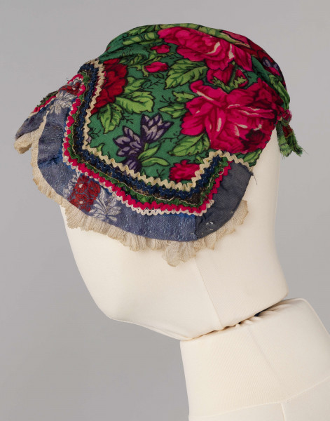 E/8141/ML - Czepek mężatki na płóciennej podszewce z zielonej cienkiej wełenki w amarantowe róże, fioletowe sasanki i jasnozielone listki. Marszczony na środku przodu i tyłu. Przód ozdobionymi kremowymi i granatowymi ząbkami, srebrną metaliczną wstążką oraz szeroką atłasową wstążką w kolorze granatowym w białe i czerwone kwiaty. Brzeg wykończony białą tiulową, drobno plisowaną kryzą. Przód wycięty w duży ząb w kształt płatka kwiatu.