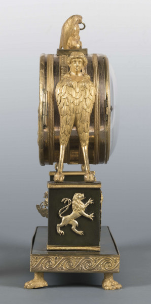 S/Mt/640/ML - Wsparty na czterech zwierzęcych łapach z brązu złoconego, podstawa prostokątna, zdobiona zgeometryzowanym fryzem, wyżej dwa prostokątne filary przechodzące w hybrydyczne figury podtrzymujące cylindryczną puszkę mechanizmu, zwieńczoną figurą orła. Tarcza emaliowana z podziałem arabskim - na niej sygnatura kursywą 