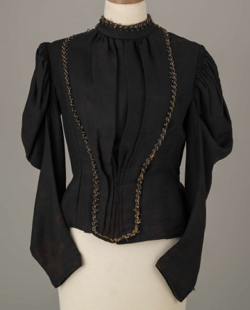 E/8038/ML - Kaftan kobiecy zszyty z 10 kawałków czarnej wełenki. Składa się z 2 części przodu, pleców i rękawów. Wycięcie wokół szyi wykończone stójką szer. 3,5 cm. Przód zapinany z boku na haftki. Rękawy w góry bufiaste, suto marszczone, zwężające się. Przód ułożony w zakładki. Stójka i przód ozdobiony żółtym sznureczkiem, czarną sieczką i białymi szklanymi koralikami. Szyty na maszynie.