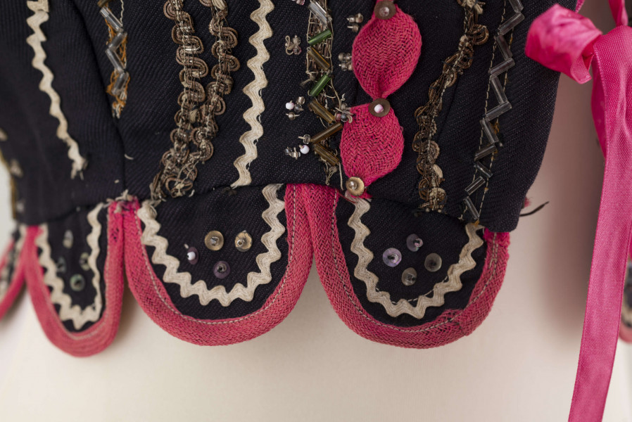 E/7358/ML - Gorset zszyty z 3 kawałków granatowej wełenki, podszyty brązową bawełnianą podszewką. Z przodu rozcięcie usztywnione fiszbinami, zaopatrzone w 10 haftek drucianych do przewlekania wstążeczki. Od pasa gorsetu dookoła opadają klapki. Cały gorset bogato zdobiony amarantowymi tasiemkami, żółtym i srebrnym szychem, białą krepiną, barwnymi koralikami i cekinami. Przód gorsetu, brzegi wykroju i pachy ozdobione dodatkowo granatowymi ząbkami. 