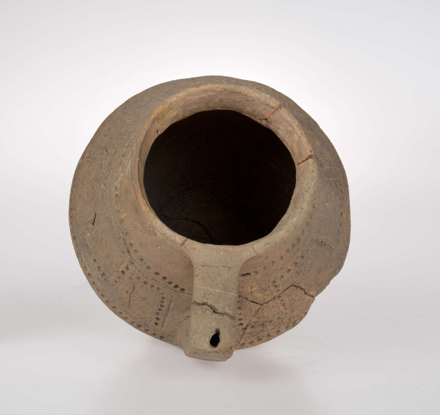 Dzban ostro profilowany, z prostym, odgiętym na zewnatrz brzegiem podkreślonym rowkiem, ostro zaznaczonym załomem brzuśca i wyodrębioną stopką. Ucho dzbana jest taśmowate, kolankowato zgięte, z przewierconym kanalikiem. Ponad załomem brzuśca naczynie zdobione jest ornamentem blankowym, który tworzy para równolegle rytych linii wypełnionych dołkami. Całość ujęta górą i dołem w wątek linii rytej i rzędu dołków. 
