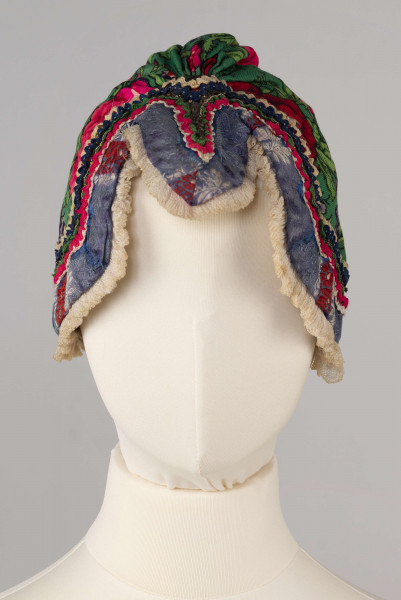 E/8141/ML - Czepek mężatki na płóciennej podszewce z zielonej cienkiej wełenki w amarantowe róże, fioletowe sasanki i jasnozielone listki. Marszczony na środku przodu i tyłu. Przód ozdobionymi kremowymi i granatowymi ząbkami, srebrną metaliczną wstążką oraz szeroką atłasową wstążką w kolorze granatowym w białe i czerwone kwiaty. Brzeg wykończony białą tiulową, drobno plisowaną kryzą. Przód wycięty w duży ząb w kształt płatka kwiatu.