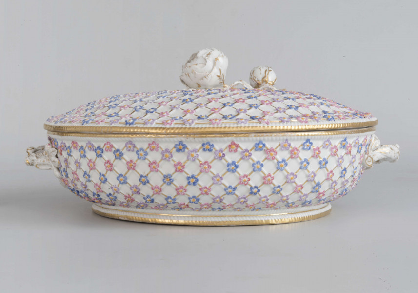 Teryna niska na pierścieniowatej podstawie, z dwoma uchami i lekko wysklepioną pokrywą. Powierzchnia zdobiona plastycznie w ukośną kratkę z drobnymi  kwiatkami (niezapominajki ?)  na przecięciach linii, brzegi karbowane. Ucha w formie gałązek z liśćmi i owocami, uchwyt pokrywy w formie pełnoplastycznego kwiatu róży z pączkiem. Brzegi, uchwyty i kratka - podkreślone złoceniami. Kwiaty malowane w trzech kolorach: błękit, róż i jasny fiolet, ich środki żółte. Wnętrze podzielone pionowymi ściankami  na trzy części. Spód naczynia nie szkliwiony.
 