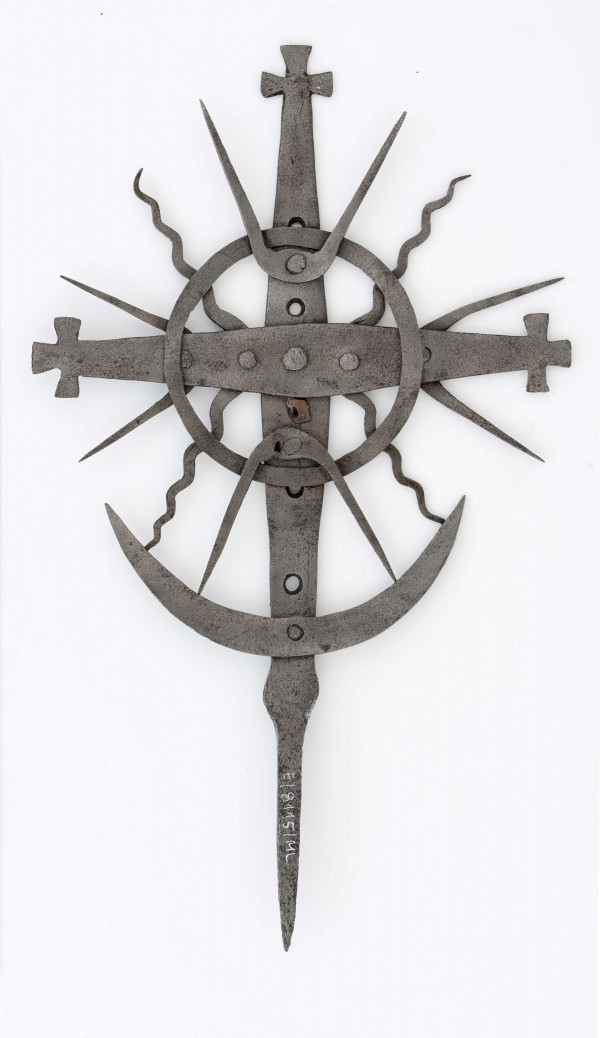 Krzyż żelazny kowalskiej roboty, stanowił element dekoracyjny przydrożnej figury lub kapliczki. Ramiona wykonane ze sztabki, przymocowane do sztychy za pomocą nitu. Końcówki ramion zakończone małymi krzyżykami. Jako element dekoracyjny, do ramion przymocowane są: promienie faliste (po 2 na ramionach bocznych i po 1 na sztycy) oraz koło o śred. 12,5 cm, wykonane z taśmy o szer. 1 cm. Każdy z promieni ma kształt litery V, przy czym nit mocujący z ramieniem, znajduje się w miejscu zgięcia. Na sztycy , w odległości 5,5 cm do koła, przymocowany jest półksiężyc, dł. 18 cm.