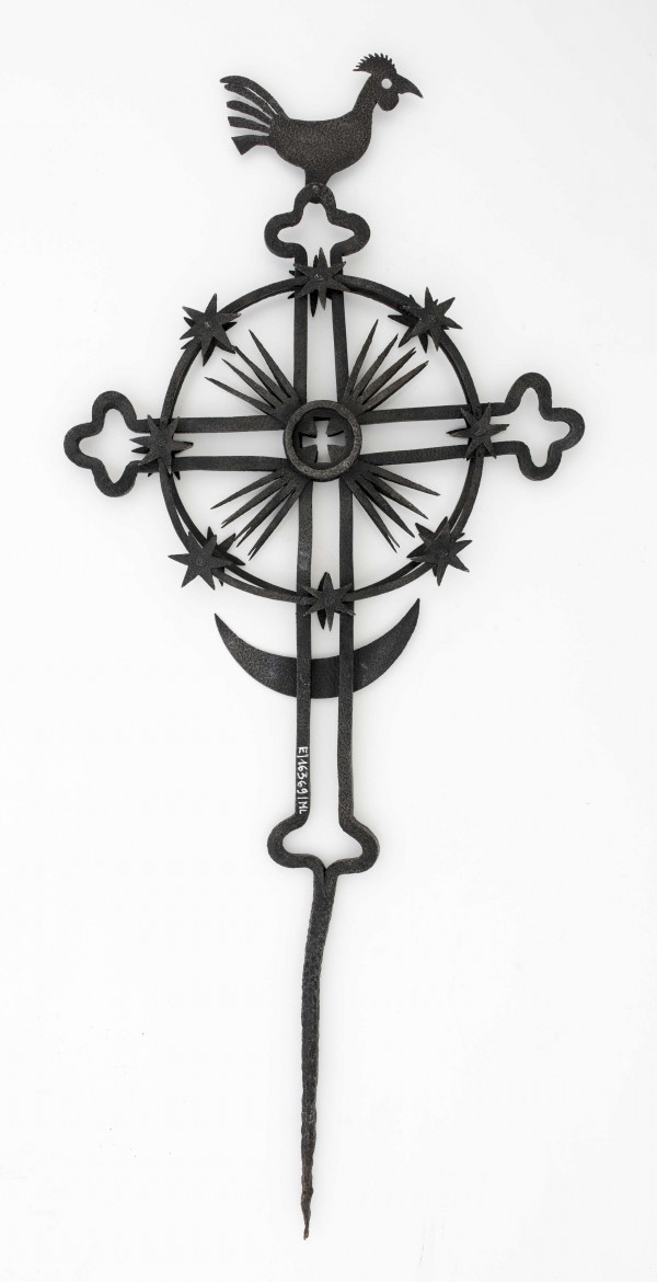 Krzyż żelazny kowalskiej roboty, stanowiący zwieńczenie drewnianego krzyża przydrożnego. Ramiona krzyża wykonane z podwójnych prętów, zakończonych na kształt listka koniczyny. Między ramionami krzyża – promienie. Ramiona z obu stron obwiedzione kołem, którego krawędź zdobią gwiazdki. U dołu krzyża półksiężyc, u góry – kogutek.