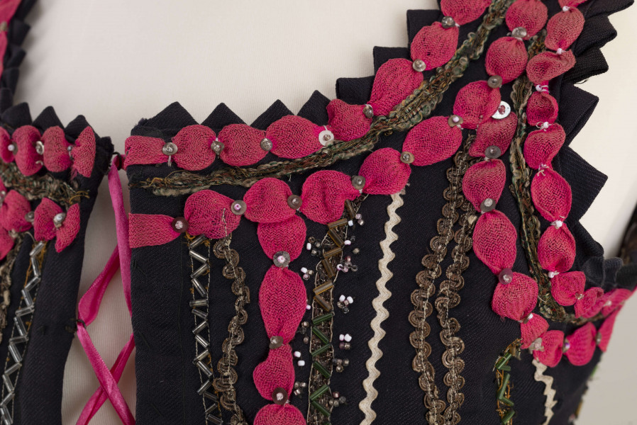 E/7358/ML - Gorset zszyty z 3 kawałków granatowej wełenki, podszyty brązową bawełnianą podszewką. Z przodu rozcięcie usztywnione fiszbinami, zaopatrzone w 10 haftek drucianych do przewlekania wstążeczki. Od pasa gorsetu dookoła opadają klapki. Cały gorset bogato zdobiony amarantowymi tasiemkami, żółtym i srebrnym szychem, białą krepiną, barwnymi koralikami i cekinami. Przód gorsetu, brzegi wykroju i pachy ozdobione dodatkowo granatowymi ząbkami. 