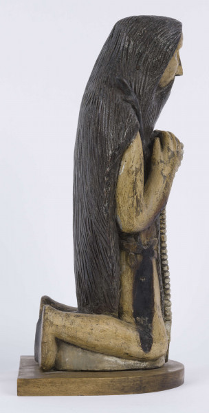 E/6353/ML - Figura drewniana, zakomponowana na postumencie, polichromowana. Postać klęcząca przedstawia św. Onufrego. Twarz płaska, trójkątna, głęboko osadzone oczy, czarne źrenice. Szerokie łuki brwiowe łączą się z wydatnym prostym nosem. Wystające kości jarzmowe. Małe usta otacza bujny zarost: broda i wąsy łączące się z bujnymi włosami, otaczającymi całą postać – na podobieństwo płaszcza. Ręce zgięte w łokciach, uniesione ku górze, łączą się końcami palców w geście modlitwy. W nieproporcjonalnie dużych dłoniach biały różaniec zakończony dużym krzyżem. Nogi masywne z dużymi płaskimi stopami. Prawą dłonią podtrzymuje sękaty kostur. W partii pasa przepasany powrozem z węzłami. 