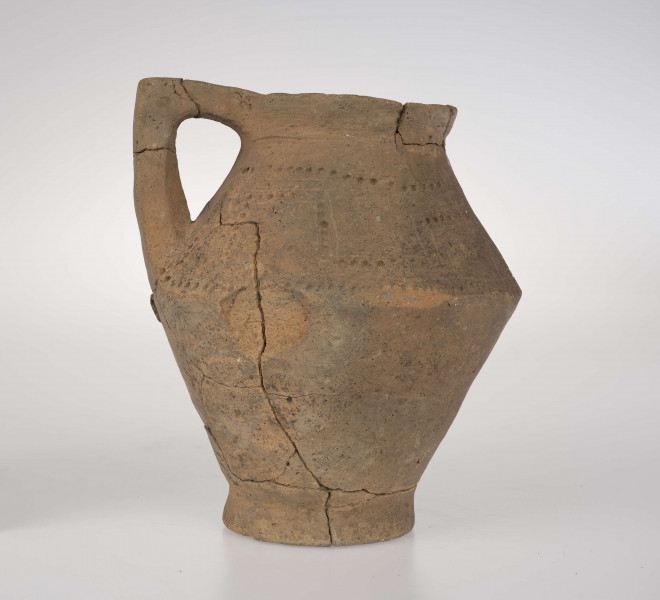 Dzban ostro profilowany, z prostym, odgiętym na zewnatrz brzegiem podkreślonym rowkiem, ostro zaznaczonym załomem brzuśca i wyodrębioną stopką. Ucho dzbana jest taśmowate, kolankowato zgięte, z przewierconym kanalikiem. Ponad załomem brzuśca naczynie zdobione jest ornamentem blankowym, który tworzy para równolegle rytych linii wypełnionych dołkami. Całość ujęta górą i dołem w wątek linii rytej i rzędu dołków. 
