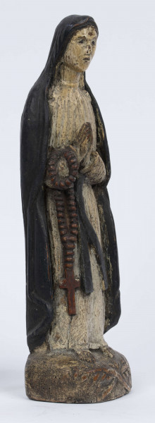 E/6363/ML - Rzeźba z drewna lipowego, polichromowana farbą olejną. Zakomponowana w jednym kawałku drewna na postumencie o formie owalnej z płaskorzeźbionym motywem dwóch skrzyżowanych gałęzi. Postać stojąca przedstawia Matkę Boską Różańcową. Twarz owalna, głęboko osadzone oczy, prosty i szeroki nos, małe pełne usta. Ręce zgięte w łokciach: duże dłonie złożone do modlitwy. Na przedramieniu prawej ręki brązowy różaniec o płaskich paciorkach, zakończony dużym krzyżem. Postać odziana w białą prostą suknię, gładko wykończoną pod szyją i przewiązaną granatową szarfą. Na głowie granatowa szata spełniająca jednocześnie rolę peleryny spływającej z ramion ku dołowi i spowijającej całą postać.