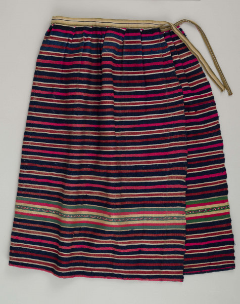 E/7351/ML - Zapaska wełniana w granatowe, czerwone, białe i amarantowe paski biegnące w poprzek. Zszyta z dwóch części – pionowy szew na przodzie. Dołem ozdobiona szerokim szlakiem dekoracyjnym, złożonym z pasków: zielonych, żółtych i amarantowych. W pasie marszczona i ujęta w oszewkę z innego materiału.