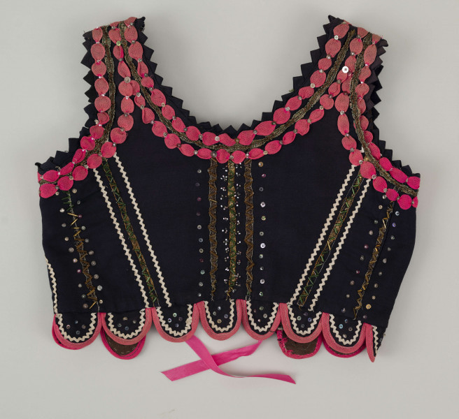 E/7358/ML - Gorset zszyty z 3 kawałków granatowej wełenki, podszyty brązową bawełnianą podszewką. Z przodu rozcięcie usztywnione fiszbinami, zaopatrzone w 10 haftek drucianych do przewlekania wstążeczki. Od pasa gorsetu dookoła opadają klapki. Cały gorset bogato zdobiony amarantowymi tasiemkami, żółtym i srebrnym szychem, białą krepiną, barwnymi koralikami i cekinami. Przód gorsetu, brzegi wykroju i pachy ozdobione dodatkowo granatowymi ząbkami. 