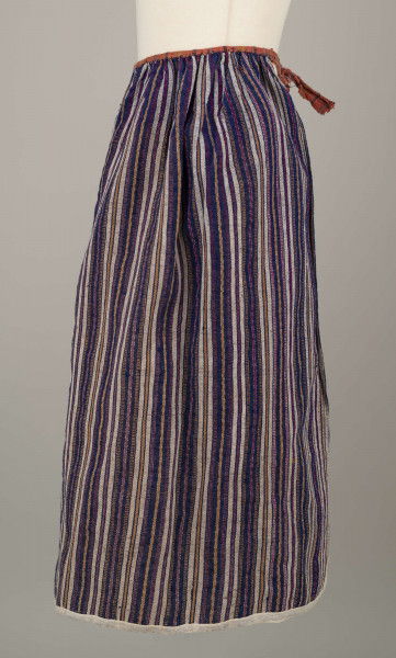 E/5997/ML - Fartuszek uszyty z jednego kawałka materiału - tkaniny samodziałowej bawełniano-wełnianej, tkanej splotem rypsowym w drobne paseczki w kolorze: białym, czerwonym, granatowym i bordo. Suto marszczony w oszewkę przechodzącą w troczki. Oszewka z materiału lnianego w kratkę czerwono-zielono-granatową. Dół obszyty tasiemką z białego bawełnianego płótna.