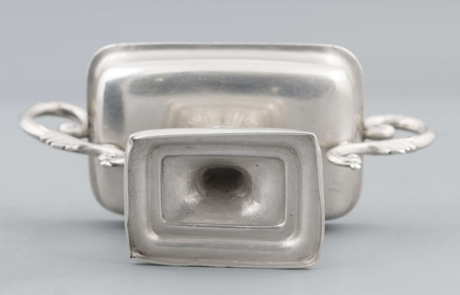 S/Mt/57/ML - Stopa prostokątna, wysklepiona, przechodząca w trzon z owalnym kapturkiem sztancowanym we wzór akantu. Czarka prostokatna o zaokrąglonych narożach i wywinietym brzegu, z dwoma uchami na krótszych bokach. Ucha wolutowe zdobione listkami. 