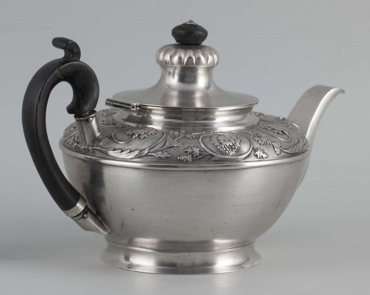 S/Mt/542/ML - Korpus w kształcie spłaszczonej kuli, na niskiej stopie, wylew w formie dzióbka, ucho drewniane malowane na czarno, osadzone w dwóch tulejkach, pokrywka na zawiasie, płaska, w części środkowej wypiętrzona i ozdobiona puklowaną rozetą.Górna część brzuśca ozdobiona sztancowaną taśmą z wzorem spiralnie zwinietej wici roślinnej z kwiatami, uchwyt przykrywki zakończony drewnianą rozetką.  
