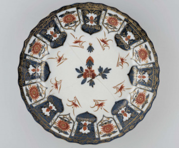 Półmisek okrągły o brzegu falistym. Na kołnierzu szeroka bordiura typu lambrekinowego z dodanymi motywami kwiatowymi, pośrodku dna gałązka kwiatowa, wokół regularnie rozmieszczone mniejsze. Dekoracja dwubarwna: partie kobaltowe malowane pod szkliwem, kwiaty malowane czerwienią naszkliwnie, całość o rysunku podkreślonym złoceniem. Na odwrocie kołnierza malowane gałęzie śliwy i irysów.
 