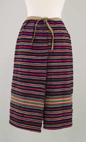 E/7351/ML - Zapaska wełniana w granatowe, czerwone, białe i amarantowe paski biegnące w poprzek. Zszyta z dwóch części – pionowy szew na przodzie. Dołem ozdobiona szerokim szlakiem dekoracyjnym, złożonym z pasków: zielonych, żółtych i amarantowych. W pasie marszczona i ujęta w oszewkę z innego materiału.