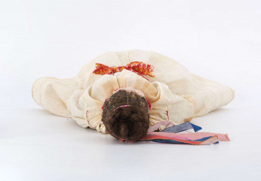 Ubiór kobiecy (model) na lalce. Składa się z bluzki haftowanej czarnymi i czerwonymi nićmi, fartucha, spódnicy i krajki. W warkocz wplecione różnokolorowe wstążki. Materiał fabryczny.