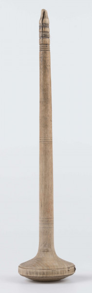 E/8019/ML - Przęślica krężołkowa z drewna brzozowego składa się z cienkiej deski – przysiadki, w której przy jednym końcu osadzona jest pionowa beleczka zw. piórem, z nasadzonym od góry ruchomym krężlem w kształcie wydłużonego stożka. Deseczka przysiadki o kształcie prostokątnym lekko zwężającym, od ok. połowy swej długości w kierunku pióra, i zakończonym głową zwierzęcą – psa. Pióro i krężel ozdobnie toczone. Krężel ma kształt stożka z rozszerzoną podstawą, w której znajduje się stożkowaty otwór, do osadzenia krężla na piórze. Dekoracja snycerska ryta znajduje się na zwężającej części przysiadki. Część zdobiona oddzielona małym stopniem. Grubość przysiadki w części zdobionej 2x większa od pozostałej części. Dekoracja snycerska tworzy ornament roślinny w postaci trzech ulistnionych drobno gałązek. Na zakończeniu ornamentu znajdują się ryte liście oraz inicjały „K.F.”. Głowa psa zdobiona drobnymi nacięcia. Zakończenie przysiadki wycięte faliście.