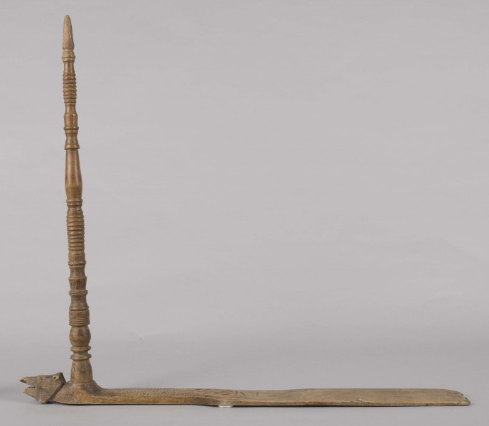 E/8019/ML - Przęślica krężołkowa z drewna brzozowego składa się z cienkiej deski – przysiadki, w której przy jednym końcu osadzona jest pionowa beleczka zw. piórem, z nasadzonym od góry ruchomym krężlem w kształcie wydłużonego stożka. Deseczka przysiadki o kształcie prostokątnym lekko zwężającym, od ok. połowy swej długości w kierunku pióra, i zakończonym głową zwierzęcą – psa. Pióro i krężel ozdobnie toczone. Krężel ma kształt stożka z rozszerzoną podstawą, w której znajduje się stożkowaty otwór, do osadzenia krężla na piórze. Dekoracja snycerska ryta znajduje się na zwężającej części przysiadki. Część zdobiona oddzielona małym stopniem. Grubość przysiadki w części zdobionej 2x większa od pozostałej części. Dekoracja snycerska tworzy ornament roślinny w postaci trzech ulistnionych drobno gałązek. Na zakończeniu ornamentu znajdują się ryte liście oraz inicjały „K.F.”. Głowa psa zdobiona drobnymi nacięcia. Zakończenie przysiadki wycięte faliście.