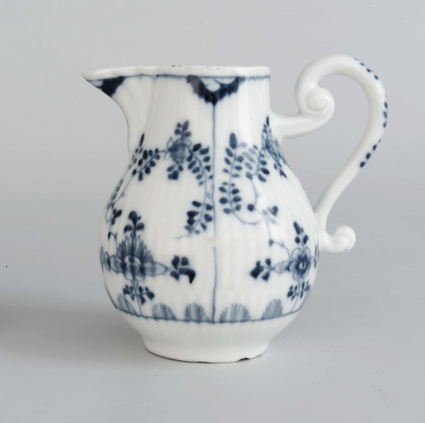 Dzbanek gruszkowaty na niskiej pierścieniowatej podstawie, ucho wolutowe, wylew trókątny, powierzchnia drobno kanelowana. Malowany podszkliwnie błękitem kobaltowym we wzór suchych kwiatów (kocanka), poniżej brzegu górnego na zewnatrz i wewnątrz malowana cienka obwódka, na dnie brzegu uszka gałązka. 
