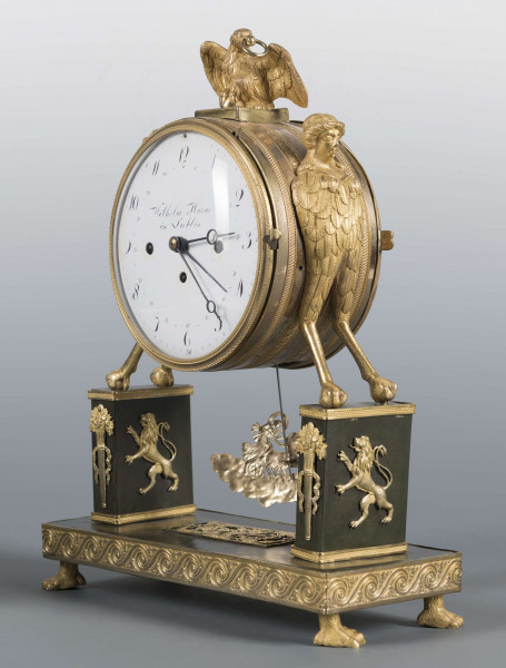 S/Mt/640/ML - Wsparty na czterech zwierzęcych łapach z brązu złoconego, podstawa prostokątna, zdobiona zgeometryzowanym fryzem, wyżej dwa prostokątne filary przechodzące w hybrydyczne figury podtrzymujące cylindryczną puszkę mechanizmu, zwieńczoną figurą orła. Tarcza emaliowana z podziałem arabskim - na niej sygnatura kursywą 