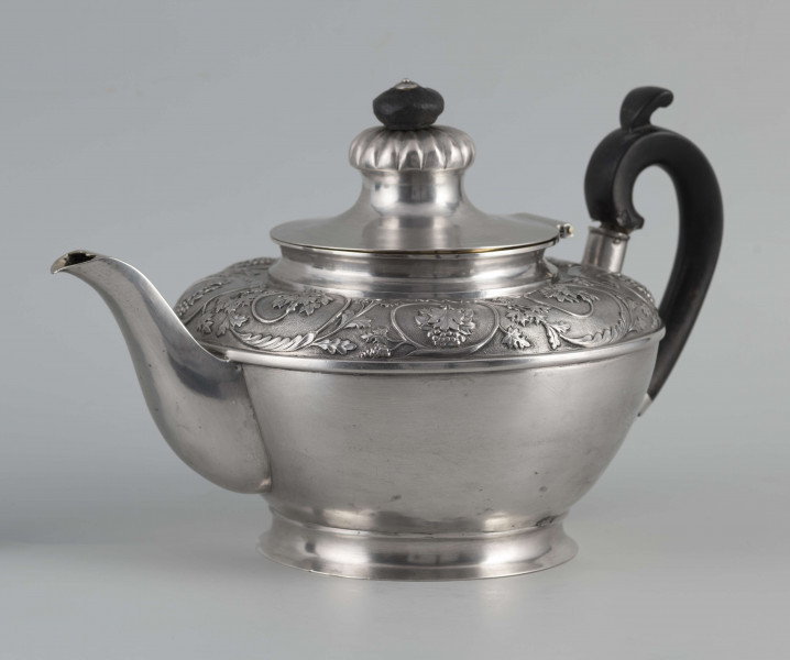 S/Mt/542/ML - Korpus w kształcie spłaszczonej kuli, na niskiej stopie, wylew w formie dzióbka, ucho drewniane malowane na czarno, osadzone w dwóch tulejkach, pokrywka na zawiasie, płaska, w części środkowej wypiętrzona i ozdobiona puklowaną rozetą.Górna część brzuśca ozdobiona sztancowaną taśmą z wzorem spiralnie zwinietej wici roślinnej z kwiatami, uchwyt przykrywki zakończony drewnianą rozetką.  
