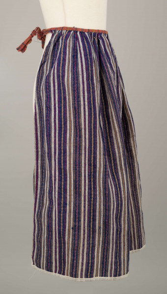 E/5997/ML - Fartuszek uszyty z jednego kawałka materiału - tkaniny samodziałowej bawełniano-wełnianej, tkanej splotem rypsowym w drobne paseczki w kolorze: białym, czerwonym, granatowym i bordo. Suto marszczony w oszewkę przechodzącą w troczki. Oszewka z materiału lnianego w kratkę czerwono-zielono-granatową. Dół obszyty tasiemką z białego bawełnianego płótna.