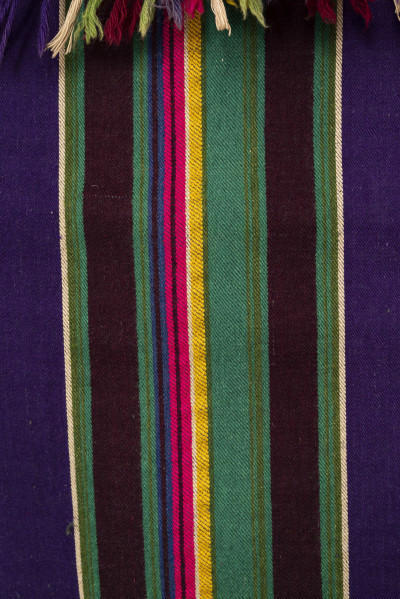 E/6045/ML - Tkanina pasiak tkana, zszyta z 2 części pośrodku. Splot rządkowym. Osnowa lniana czarna, wątek wełniany w kolorach: fioletowy, zielony, brązowy, żółty, amarantowy, biały, ciemnoniebieski. Raport tworzą poprzeczne wiązki pasów oddzielone szerokimi pasami w kolorze fioletowym. Jedna z krawędzi wzdłużnych wykończona szydełkową koronką z wełny w kolorach występujących w tkaninie. Zakończona wiązanymi frędzlami. 