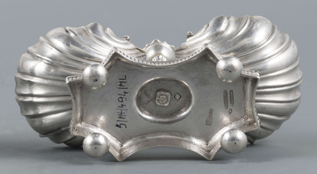 S/Mt/494/ML - Solniczka składa się z dwóch czarek w formie muszli połączonych z pionowym, profilowanym uchwytem o zwieńczniu w formie makówki ustawionym na wydłużonej ośmiobocznej podstawie o wklęsłych bokach. Krawędź podstawy fasetowana, boki karbowane, cztery nóżki w kształcie spłaszczonych kulek. Wnetrze muszli złocone.
Na dnie podstawy sygnatura kursywą w owalnych ramkach:
- J.Gałecki
- w Lublinie
- punca owalna z przedstawieniem dłoni (sygnatura pomocnicza)
- wężyk probierczy
- znak probierni warszawskiej z próbą 