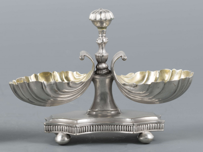 S/Mt/494/ML - Solniczka składa się z dwóch czarek w formie muszli połączonych z pionowym, profilowanym uchwytem o zwieńczniu w formie makówki ustawionym na wydłużonej ośmiobocznej podstawie o wklęsłych bokach. Krawędź podstawy fasetowana, boki karbowane, cztery nóżki w kształcie spłaszczonych kulek. Wnetrze muszli złocone.
Na dnie podstawy sygnatura kursywą w owalnych ramkach:
- J.Gałecki
- w Lublinie
- punca owalna z przedstawieniem dłoni (sygnatura pomocnicza)
- wężyk probierczy
- znak probierni warszawskiej z próbą 