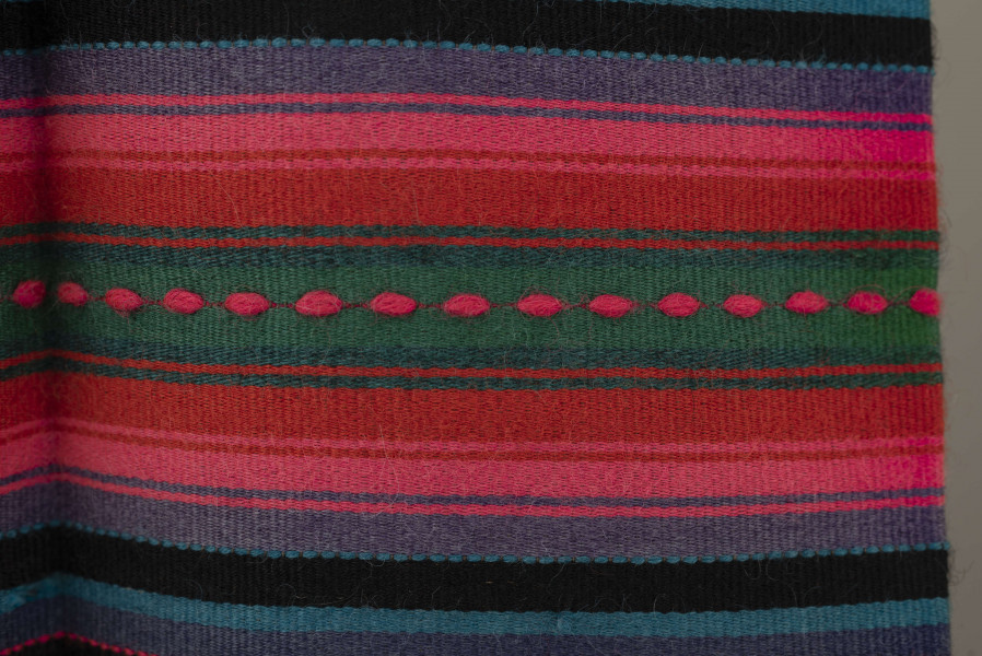 E/6813/ML - Fartuszek wełniany tkany w kolorowe wąskie pasy w kolorze: granatowym, niebieskim, fioletowym, czarnym i amarantowym z szerokim pasem o wiązce kolorystycznej: amarantowy, czerwony, zielony. Przymarszczony i wszyty w płócienny pasek z troczkami.