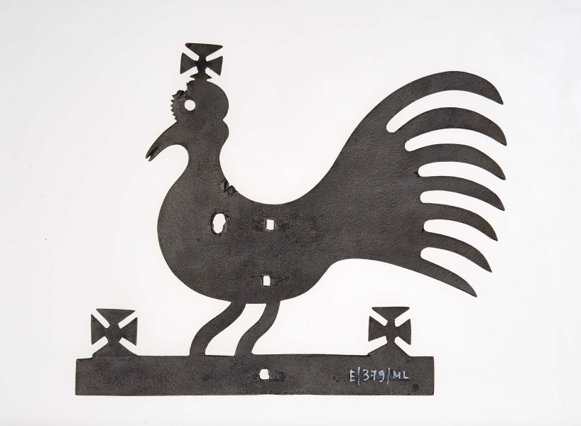 Kogut wykonany z żelaza techniką kucia. Stanowi zakończenie żelaznego krzyża zwieńczającego drewniany krzyż przydrożny. Posiada krzyżyki na głowie i w obu końcach podstawy.
