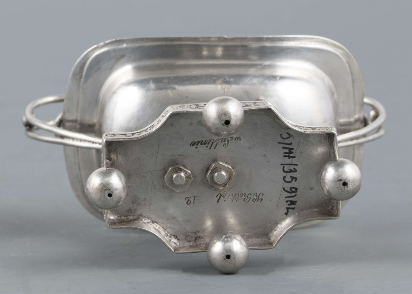 S/Mt/359/ML - Solniczka w kształcie prostokątnej łódki z dwoma wolutowymi uchami z okrągłego drutu, umieszczonej na figurce gryfa, z  podstawą prostokątną o wklęsłych narożach i czterech nóżkach w formie spłaszczonych kulek. Brzeg łódki wywinięty, wnętrze złocone. Brzegi łódki i boki podstawy obiega moletowana taśma z ornamentem roślinnym.