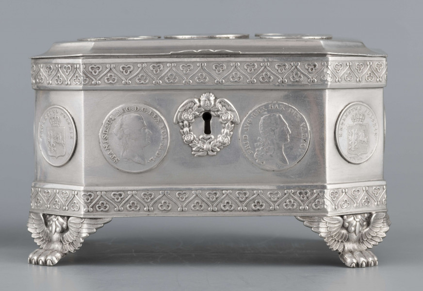 S/Mt/364/ML - Cukiernica skrzynkowa prostokątna ze ściętymi narożami, na czterech nóżkach w formie uskrzydlonych lwich łap, z wieczkiem o wklęsło profilowanym brzegu. Górny i dolny brzeg obiega  moletowana taśma z motywem neogotyckich trójliści, szyld zamka ma formę różanego wieńca pod skrzydatą główką..W ścianki i wieczko wmontowanych jest 15 monet srebrnych:
1 ort Zygmunta III
1 talar pruski, 1818 r.
1 pięciofrankówka, 1811 r.
1 moneta austriacka, 1797 r.
1 moneta saska, 
2 monety polskie St.Augusta Poniatowskiego
1 dwózłotówka Królestwa Polskiego, 1825 r.
1 pięciozłotówka, 1831 r.
6 złotówek, 1831 r.
Na dnie sygnatura wklęsła kursywą:
- K. Rotkiel w Lublinie
i punca z oznaczeniem próby 