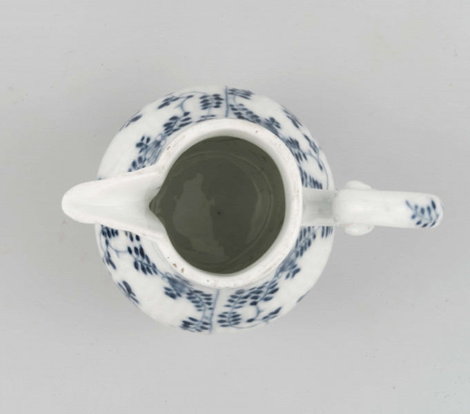 Dzbanek gruszkowaty na niskiej pierścieniowatej podstawie, ucho wolutowe, wylew trókątny, powierzchnia drobno kanelowana. Malowany podszkliwnie błękitem kobaltowym we wzór suchych kwiatów (kocanka), poniżej brzegu górnego na zewnatrz i wewnątrz malowana cienka obwódka, na dnie brzegu uszka gałązka. 
