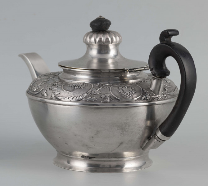 S/Mt/542/ML - Korpus w kształcie spłaszczonej kuli, na niskiej stopie, wylew w formie dzióbka, ucho drewniane malowane na czarno, osadzone w dwóch tulejkach, pokrywka na zawiasie, płaska, w części środkowej wypiętrzona i ozdobiona puklowaną rozetą.Górna część brzuśca ozdobiona sztancowaną taśmą z wzorem spiralnie zwinietej wici roślinnej z kwiatami, uchwyt przykrywki zakończony drewnianą rozetką.  
